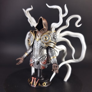 Peut inclure: Buste d&eacute;taill&eacute; d'une figurine en armure argent&eacute;e et dor&eacute;e avec une cape &agrave; capuche brune. La figurine est pos&eacute;e sur un socle noir avec le mot "Diablo" inscrit dessus. La figurine est entour&eacute;e de filaments blancs.