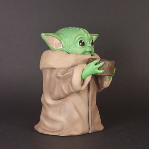 K&ouml;nnte beinhalten: Eine gr&uuml;ne und braune Figur eines Charakters aus dem Star Wars-Franchise, die eine kleine braune Tasse h&auml;lt. Der Charakter hat gro&szlig;e Ohren und einen gro&szlig;en Kopf.