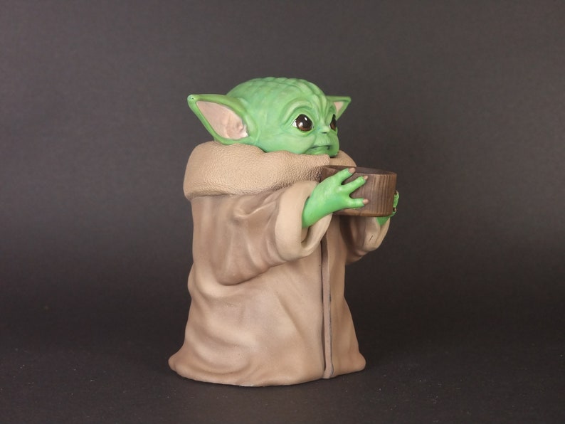K&ouml;nnte beinhalten: Eine gr&uuml;ne und braune Figur eines Charakters aus dem Star Wars-Franchise, die eine kleine braune Tasse h&auml;lt. Der Charakter hat gro&szlig;e Ohren und einen gro&szlig;en Kopf.