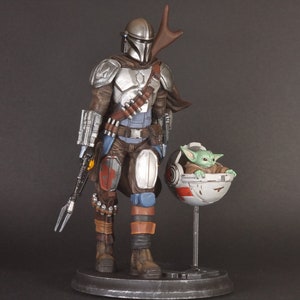 Op de afbeelding: Een zilveren en bruine Mandalorian action figure met een groen alien personage in een zwevende pod. De Mandalorian houdt een wapen vast en heeft een cape. Het alien personage zit in de pod met zijn armen uitgestrekt.