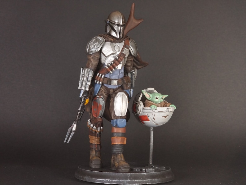 Op de afbeelding: Een zilveren en bruine Mandalorian action figure met een groen alien personage in een zwevende pod. De Mandalorian houdt een wapen vast en heeft een cape. Het alien personage zit in de pod met zijn armen uitgestrekt.
