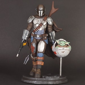 Op de afbeelding: Een zilveren en bruine Mandalorian action figure met een groen alien personage in een zwevende pod. De Mandalorian houdt een blastergeweer en een speer vast. De figuur staat op een zwarte basis.