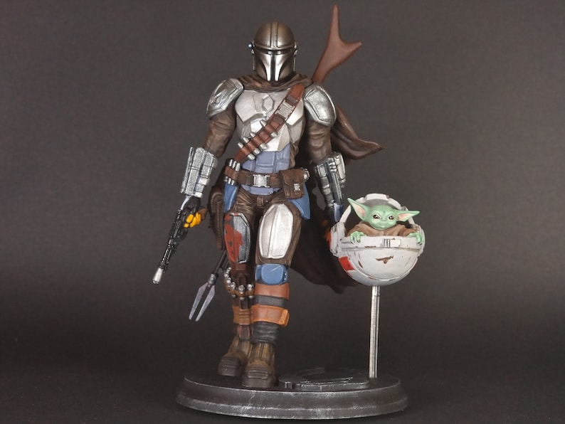 Op de afbeelding: Een zilveren en bruine Mandalorian action figure met een groen alien personage in een zwevende pod. De Mandalorian houdt een blastergeweer en een speer vast. De figuur staat op een zwarte basis.