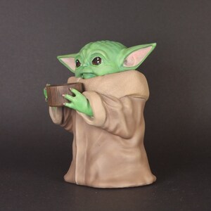 K&ouml;nnte beinhalten: Eine gr&uuml;ne und braune Figur eines Charakters aus dem Star Wars-Franchise, die eine kleine braune Sch&uuml;ssel in den H&auml;nden h&auml;lt.