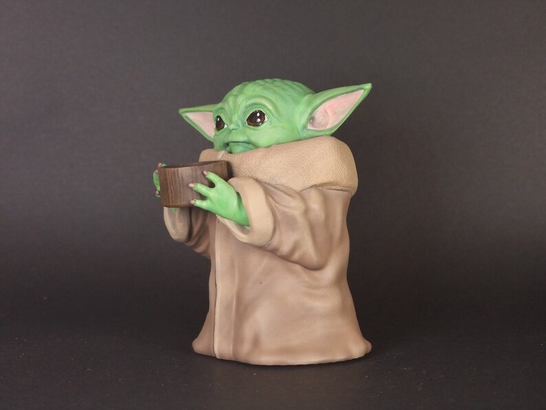 K&ouml;nnte beinhalten: Eine gr&uuml;ne und braune Figur eines Charakters aus dem Star Wars-Franchise, die eine kleine braune Sch&uuml;ssel in den H&auml;nden h&auml;lt.