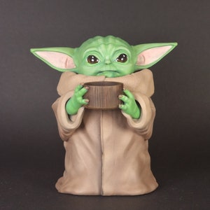 K&ouml;nnte beinhalten: Eine gr&uuml;ne und braune Figur eines Charakters aus dem Star Wars-Franchise, die ein braunes zylindrisches Objekt in den H&auml;nden h&auml;lt.