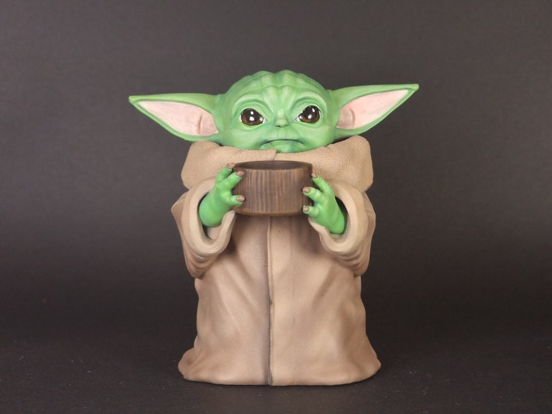 K&ouml;nnte beinhalten: Eine gr&uuml;ne und braune Figur eines Charakters aus dem Star Wars-Franchise, die ein braunes zylindrisches Objekt in den H&auml;nden h&auml;lt.