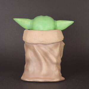 K&ouml;nnte beinhalten: Ein gr&uuml;nes und braunes Pl&uuml;schtier, das einen Charakter aus dem Star Wars-Franchise darstellt. Das Spielzeug tr&auml;gt eine braune Robe und hat gro&szlig;e Ohren.