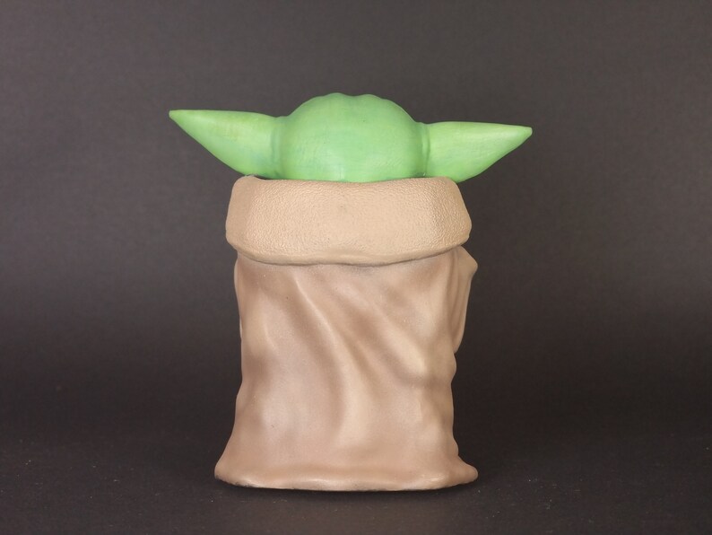 K&ouml;nnte beinhalten: Ein gr&uuml;nes und braunes Pl&uuml;schtier, das einen Charakter aus dem Star Wars-Franchise darstellt. Das Spielzeug tr&auml;gt eine braune Robe und hat gro&szlig;e Ohren.