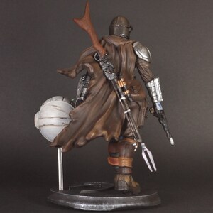 Op de afbeelding: Een gedetailleerd actiefiguur van een personage met een bruine mantel en helm, dat een wapen vasthoudt en op een voetstuk staat. De figuur is gemaakt van plastic en is in een realistische stijl geschilderd.