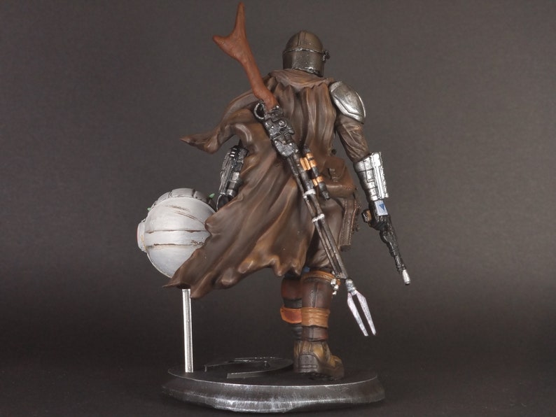Op de afbeelding: Een gedetailleerd actiefiguur van een personage met een bruine mantel en helm, dat een wapen vasthoudt en op een voetstuk staat. De figuur is gemaakt van plastic en is in een realistische stijl geschilderd.