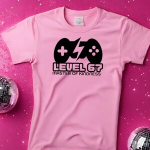 Può includere: Maglietta rosa chiaro con un disegno nero di controller di gioco e la scritta "LEVEL 67 MASTER OF KINDNESS". L'immagine include sfere da discoteca e il testo "digital download PNG + JPG" su sfondo rosa.