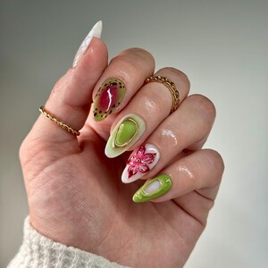 May include: Close-up of a hand with green, white, and pink nail art. The nails are long and almond shaped. The art features abstract shapes, a flower, and gold accents.