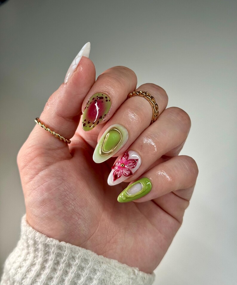 May include: Close-up of a hand with green, white, and pink nail art. The nails are long and almond shaped. The art features abstract shapes, a flower, and gold accents.