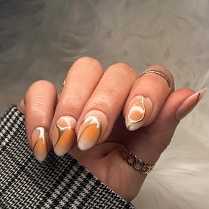 May include: Close-up of a hand with almond-shaped nails painted with a gradient of white and orange. The nails have a gold abstract design.