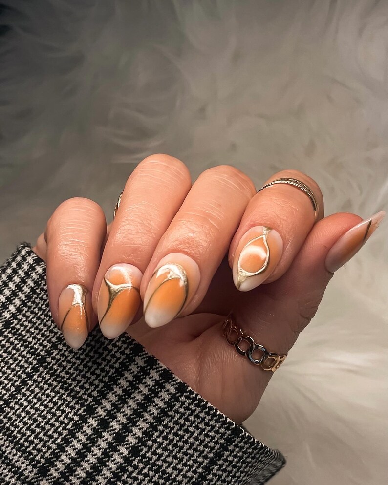May include: Close-up of a hand with almond-shaped nails painted with a gradient of white and orange. The nails have a gold abstract design.