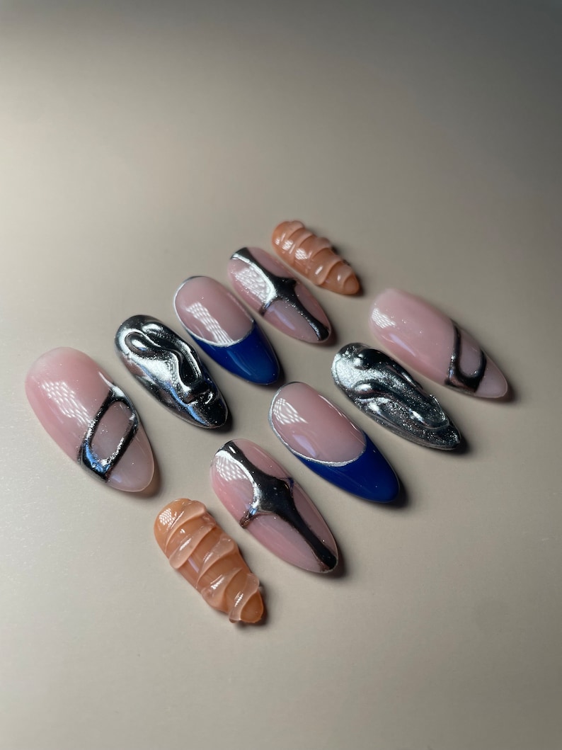 May include: Set of eight false nails with a nude base and silver and blue accents. The nails feature a variety of designs, including swirls, lines, and geometric shapes.