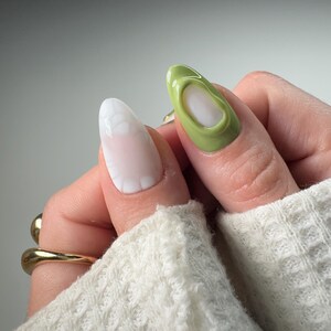 May include: Close-up of two fingernails with different nail art designs. One nail is painted a light green color with a 3D design, while the other nail is painted white with a white 3D design.