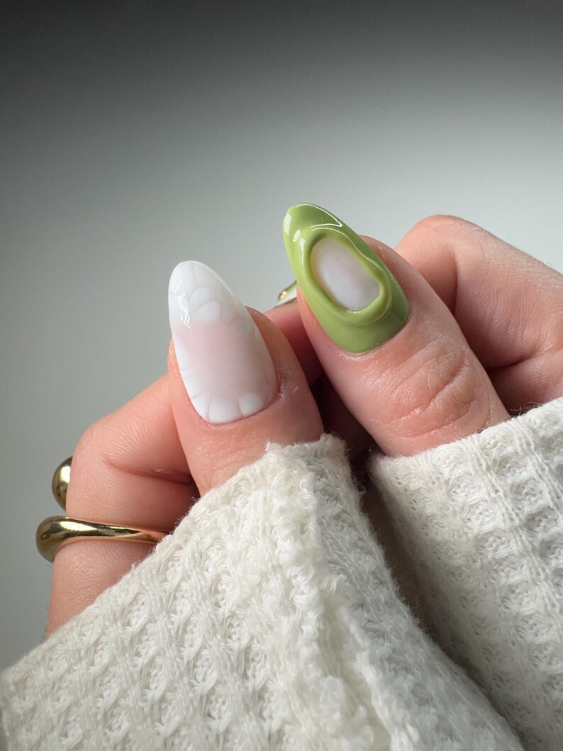 May include: Close-up of two fingernails with different nail art designs. One nail is painted a light green color with a 3D design, while the other nail is painted white with a white 3D design.
