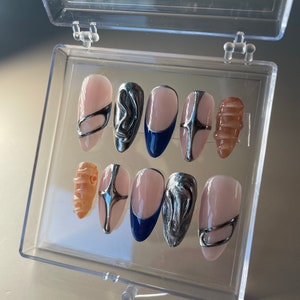 May include: A set of ten false nails in a clear plastic box. The nails are oval shaped and have a light pink base. Some nails have silver abstract designs, others have a silver band, and one has a blue tip.