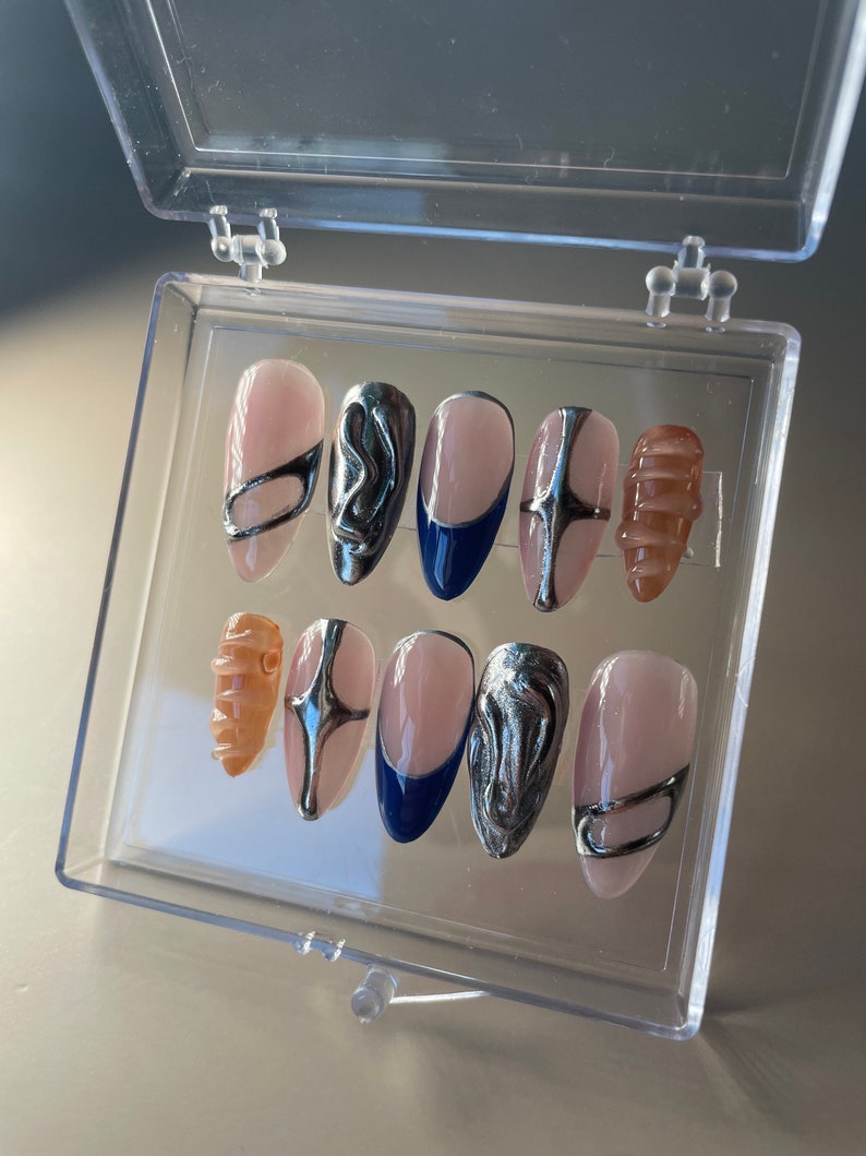 May include: A set of ten false nails in a clear plastic box. The nails are oval shaped and have a light pink base. Some nails have silver abstract designs, others have a silver band, and one has a blue tip.