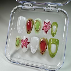 May include: A set of ten false nails in a clear plastic container. The nails are decorated with different designs, including white, green, red, and gold. Some nails have a flower design, while others have a polka dot design.