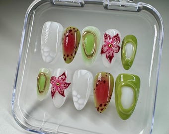 May include: A set of ten false nails in a clear plastic container. The nails are decorated with different designs, including white, green, red, and gold. Some nails have a flower design, while others have a polka dot design.