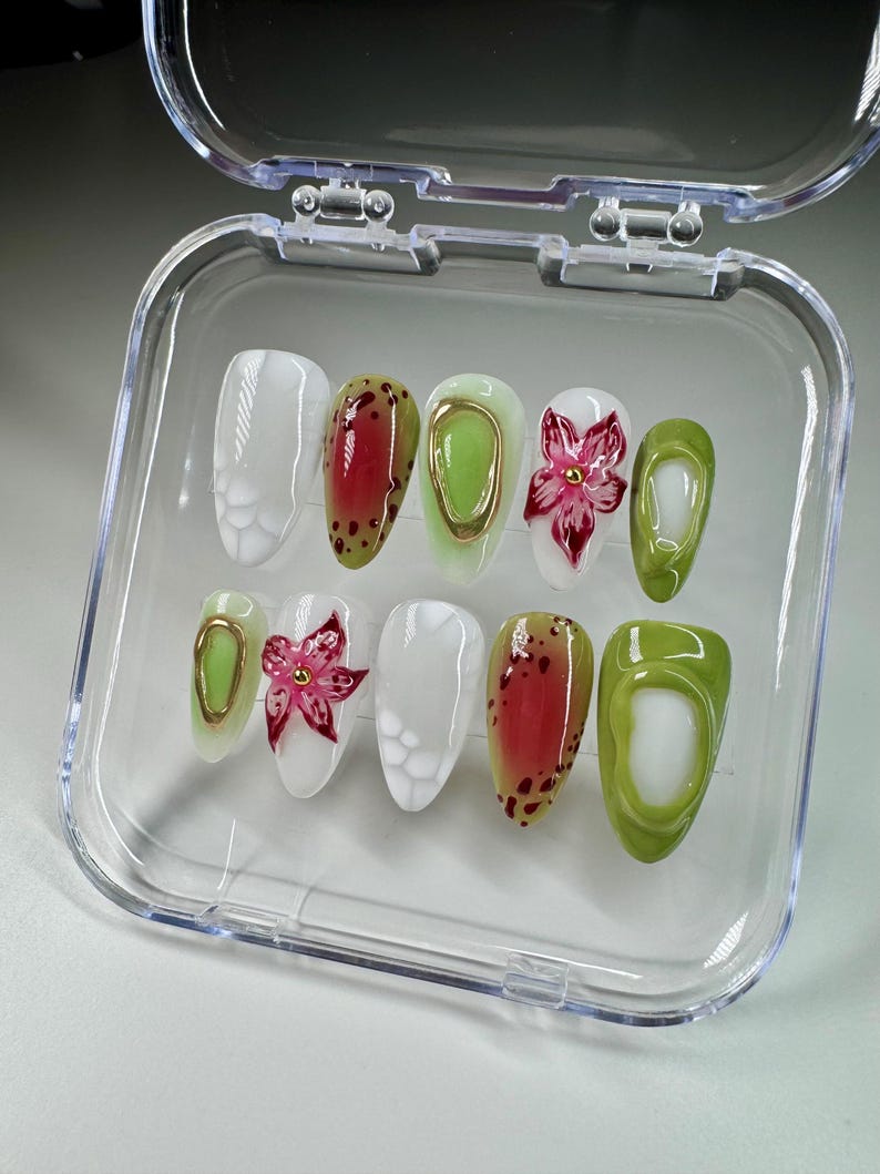 May include: A set of ten false nails in a clear plastic container. The nails are decorated with different designs, including white, green, red, and gold. Some nails have a flower design, while others have a polka dot design.