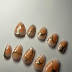 May include: Set of ten peach-colored almond-shaped press-on nails with gold abstract designs.