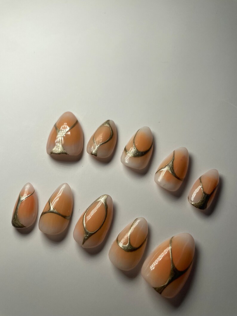 May include: Set of ten peach-colored almond-shaped press-on nails with gold abstract designs.