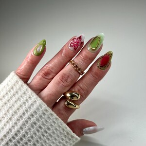 May include: Close-up of a hand with green, white, and red nail polish. The nails are decorated with flowers, gold accents, and a red and white design. The hand is wearing two gold rings.