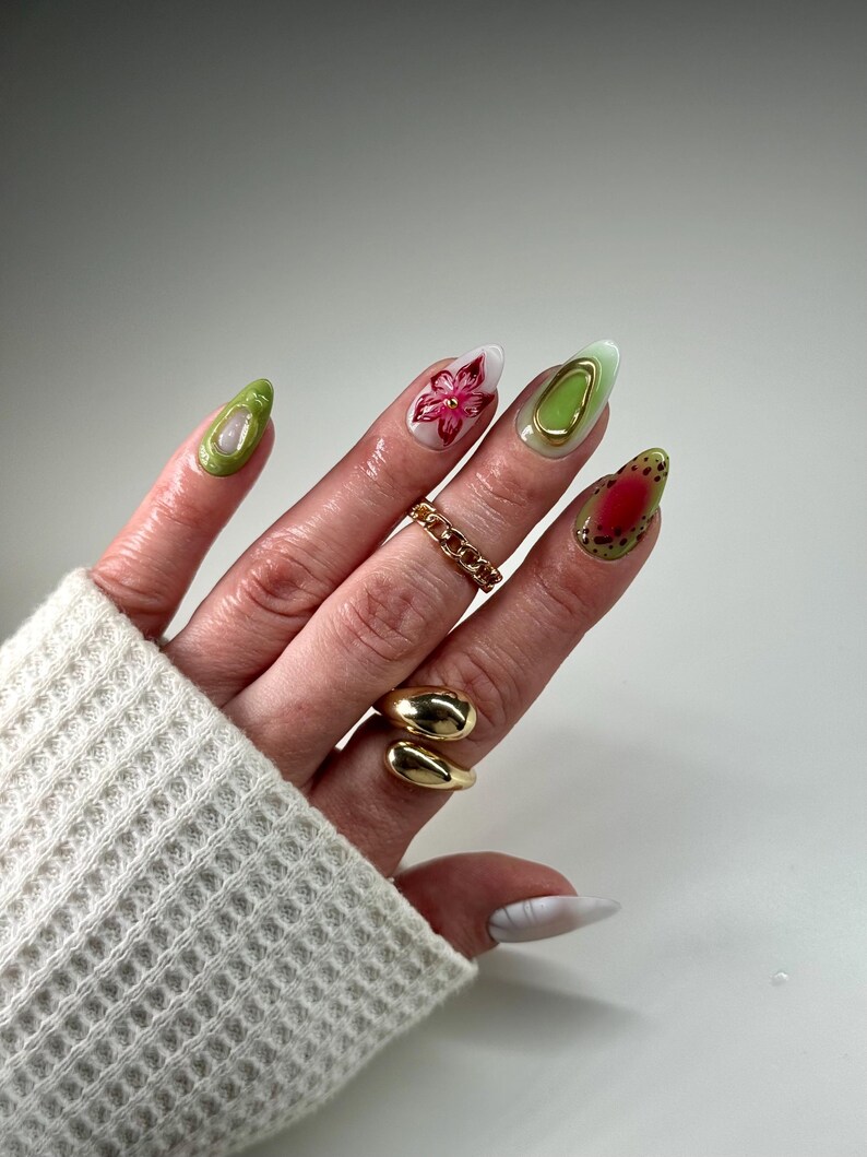 May include: Close-up of a hand with green, white, and red nail polish. The nails are decorated with flowers, gold accents, and a red and white design. The hand is wearing two gold rings.