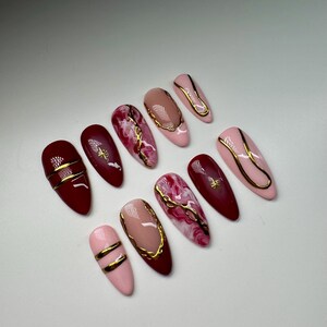 May include: Set of ten false nails with a burgundy and pink base and gold accents. Some nails have a marble design, while others have a simple gold line design.