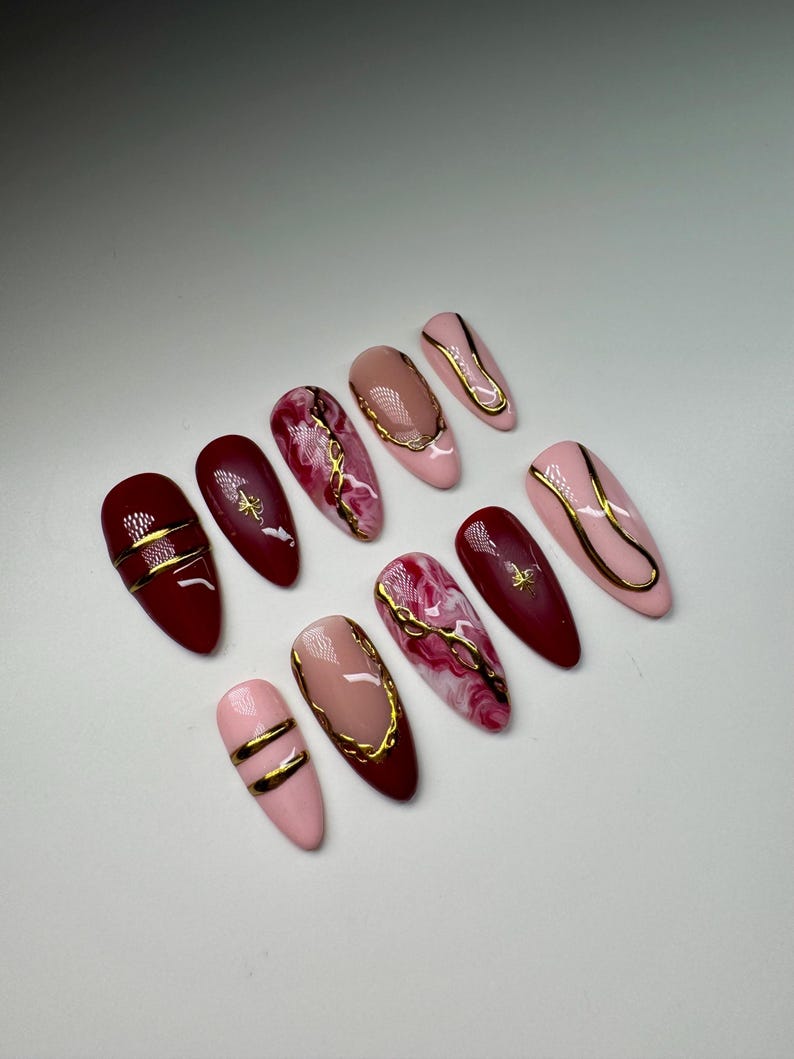 May include: Set of ten false nails with a burgundy and pink base and gold accents. Some nails have a marble design, while others have a simple gold line design.