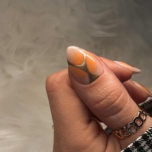 May include: A close-up of a finger with a light orange and white nail polish design. The nail is shaped into an almond shape and has a gold geometric design on top. The finger is wearing a gold chain ring.