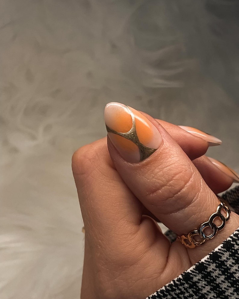 May include: A close-up of a finger with a light orange and white nail polish design. The nail is shaped into an almond shape and has a gold geometric design on top. The finger is wearing a gold chain ring.