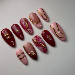 May include: Set of ten false nails with a burgundy and pink base and gold accents. The nails have a variety of designs, including swirls, lines, and geometric shapes.