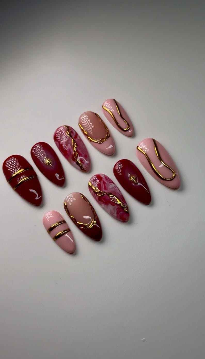 May include: Set of ten false nails with a burgundy and pink base and gold accents. The nails have a variety of designs, including swirls, lines, and geometric shapes.