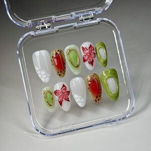 May include: A set of ten false nails in a clear plastic case. The nails are oval shaped and have different designs. Some nails are white with a small flower design, some are green, some are red with black spots, and some have a gold accent.