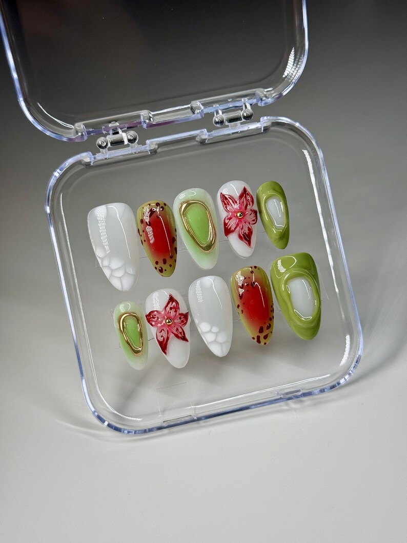 May include: A set of ten false nails in a clear plastic case. The nails are oval shaped and have different designs. Some nails are white with a small flower design, some are green, some are red with black spots, and some have a gold accent.
