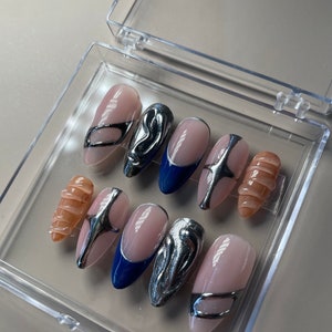 May include: A set of ten false nails in a clear plastic box. The nails are oval shaped and have a variety of designs. Some nails are nude with silver accents, some are blue with silver accents, and some are nude with a swirl design.