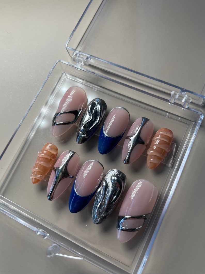 May include: A set of ten false nails in a clear plastic box. The nails are oval shaped and have a variety of designs. Some nails are nude with silver accents, some are blue with silver accents, and some are nude with a swirl design.