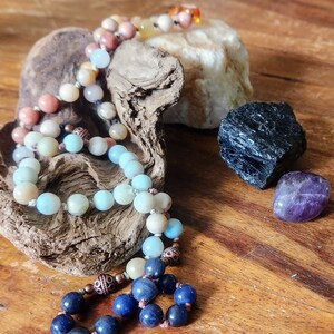 May include: A multi-colored beaded necklace with blue, pink, and white beads. The necklace is resting on a piece of driftwood with a black stone and a purple amethyst stone nearby.