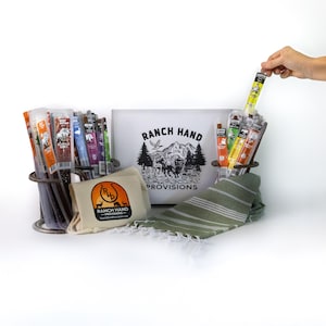 May include: A gift set featuring a variety of beef jerky sticks in a metal holder, a small canvas bag with the Ranch Hand Provisions logo, a green and white striped towel, and a white sign with the text "Ranch Hand Provisions" and a mountain scene.