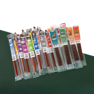 May include: An assortment of individually packaged meat sticks, featuring various flavors and animal types. The colorful packaging includes text and illustrations of animals like elk, alligator, and wild boar. The sticks are displayed on a dark green surface.