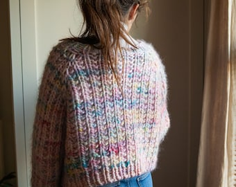 May include: A chunky knit sweater in a light pink hue with colorful flecks of blue, yellow, and pink. The sweater has a ribbed texture and a cropped fit. The model is wearing blue jeans. The sweater is a cozy and stylish garment.