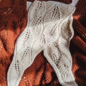May include: A white knitted scarf with a leaf pattern, laid on a brown knitted blanket.