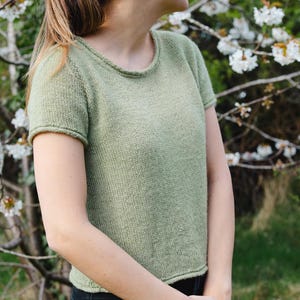 以下が含まれることがあります： 丸首の緑色のニットの半袖トップスを着た女性。白い花が咲いた木の前に立っている。