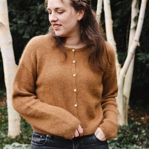 Könnte beinhalten: Ein brauner Cardigan mit Perlmuttknöpfen, kombiniert mit dunkelblauen Jeans. Das Bild wurde im Freien mit Bäumen im Hintergrund aufgenommen. Der Cardigan hat einen runden Ausschnitt und lange Ärmel. Ein warmes Strickkleidungsstück.