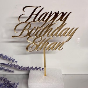 May include: A gold-colored cake topper with the words "Happy Birthday Ethan" in elegant script. The topper is mounted on a slender gold stem and a white foam base. Lavender sprigs are in the background.